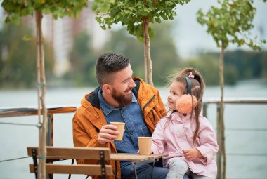 Dışarıda bir kafede çocukla vakit geçirirken elinde kahve tutan ve gülümseyen bir adam. Mutlu bir baba ve kızı, New Kentsel Bölgesi 'nin caddesinde masada oturuyor. Erkek ve küçük kıza odaklan..