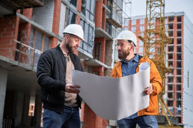 Kasklı sakallı adam elinde mimari planla dışarıda inşaat işçisiyle konuşuyor. İnşaat alanında uzmanlaşmış bir erkek ev sahibi tartışma binası planı.
