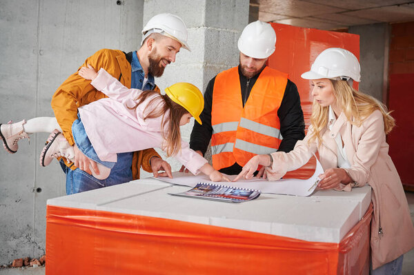 Woman standing next to builder and pointing at building plan while man holding daughter. Family with child discussing architectural plan of new apartment with construction worker.