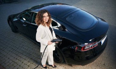 Güzel bir iş kadınının kameraya bakıp elektrikli araba şarj aletini tutarken gülümsemesinin en üst görüntüsü. Beyaz takım elbiseli şık bir kadın araç şarj istasyonunda elektrikli arabanın yanında duruyor..