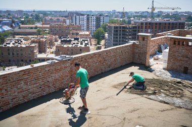 Konut binasının çatısını tararken troweling makinesi kullanan bir adam. Yeni binadaki zemini tararken düz kenarlı başka bir adam yüzeyi düzleştiriyor..