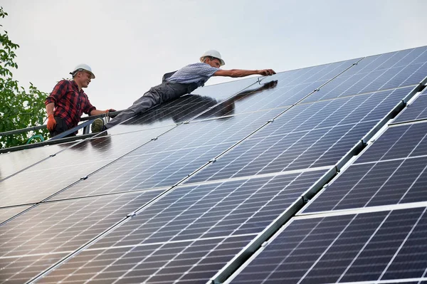 Male workers mounting solar modules for generating electricity through photovoltaic effect. Two men solar technicians in safety helmets assembling solar panels under blue white sky.