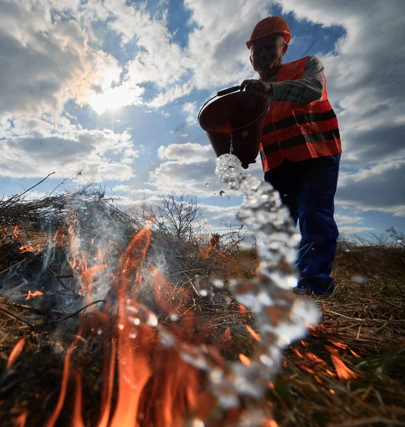 Fireman ecologist extinguishing fire in field with cloudy sky on background. Low angle view male environmentalist holding bucket and pouring water on burning dry grass. Natural disaster concept.