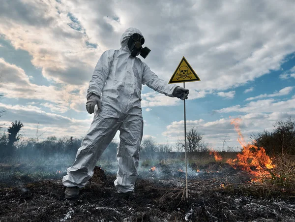 Firefighter ecologist extinguishing fire in field. Man in suit and gas mask near burning grass with smoke, holding yellow triangle with skull and crossbones warning sign. Natural disaster concept.