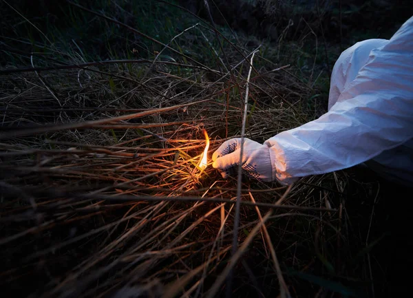 Man burning old dried grass in field. Close up of hand holding burning match and setting fire to dry grass at night. Concept of ecology and human factor in fires.