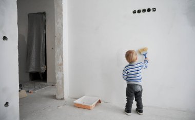 Back view of little boy painting white wall in empty room under renovation. Cute kid using paint brush while renovating apartment. Concept of home renovation, restoration and childhood. clipart