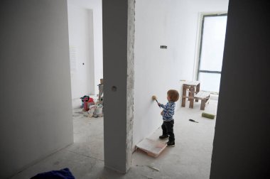 Tadilat altındaki iki oda arasında beyaz duvar boyayan sevimli bir erkek çocuk. Evini yenilerken boya fırçası kullanan sevimli bir çocuk. Ev yenileme, restorasyon ve çocukluk kavramı.