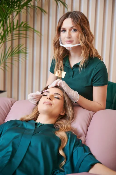 Portrait of beauty specialist comping client eyelashes after extension procedure. Female eyelashes technician in protective face mask combing woman lashes with disposable mascara brush in beauty salon