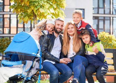 Genç ebeveynlerin bebek arabasında üç çocuk ve bebekle bankta oturup şehirde dinlenmesi ön planda. İki yetişkinin sevimli çocuklarla vakit geçirip kameraya gülümsediği portre görüntüsü. Aile kavramı.