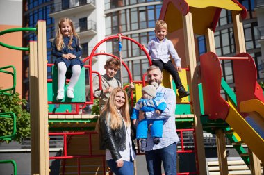 Üç sevimli çocuk anne babasıyla ve yeni doğmuş bebekle oyun bahçesinde takılıyor. Güneşli bir günde diğer çocuklar gülümserken, kucağında bebek olan mutlu anne ve babalarının ön görüntüsü. Aile kavramı.