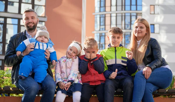 Üç çocuk ve bir bebeğin babalarının kollarında oturduğu bankta oturan genç ebeveynlerin portre görüntüsü. İki yetişkinin kameraya gülümseyen dört sevimli çocukla vakit geçirmesi. Aile kavramı.