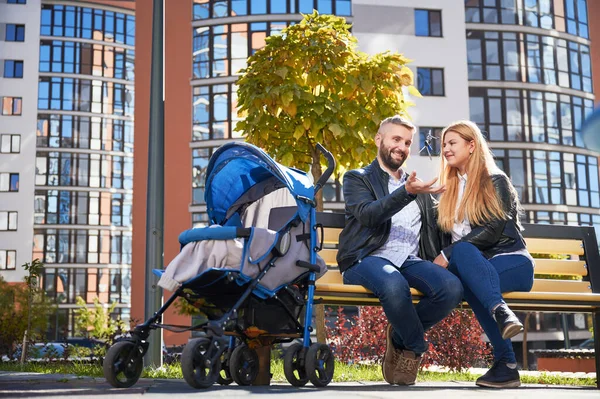 快乐的一家人坐在一起 带着蓝色婴儿车滑行 微笑的男人从新城区的新公寓里抛出钥匙 背景的现代住宅 — 图库照片