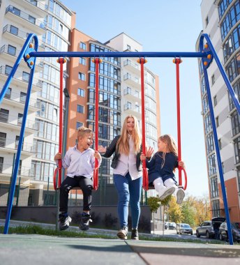 Neşeli kardeşler salıncakta sallanırken, mutlu anne çocuk parkında iterken. Şehir merkezinin modern avlusunda güzel kız ve oğlan sallayan bir kadın..