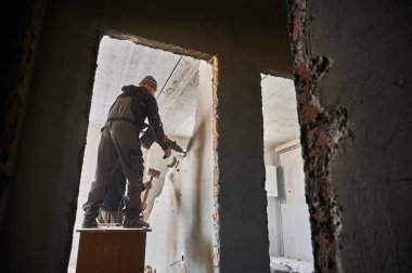 Binadaki duvarı yıkarken çekiç matkabı ve balyoz kullanan erkek inşaatçılar. Tadilat altındaki apartmanın duvarlarını yıkan iki işçinin kapı girişine bakın..