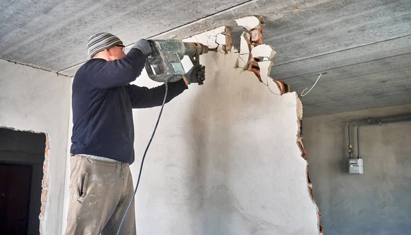 Elektrikli çekiç matkabıyla duvarı delen adam. Erkek işçi, tadilat sırasında evdeki duvarı yıkarken profesyonel sondaj aletini kullanıyor. Ev yenileme ve yeniden yapılandırma kavramı.