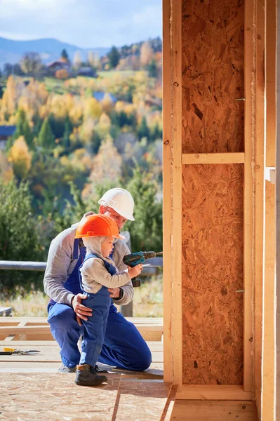 Father with toddler son building wooden frame house. Boy helping his daddy, using screwdriver on construction site, wearing helmet and blue overalls on sunny day. Carpentry and family concept.