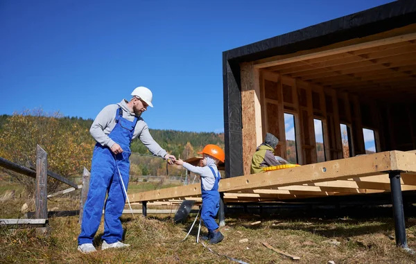Father with toddler son building wooden frame house on pile foundation. Boy helping his daddy, playing with tape measure on construction site, wearing helmet and blue overalls. Carpentry concept.