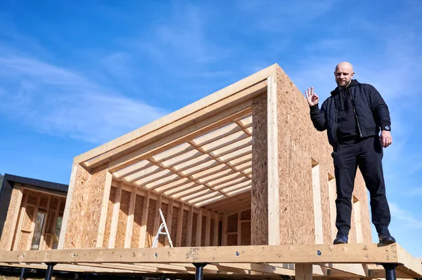 Erkek inşaatçı ahşap çerçeve inşa ediyor. İnşaat alanında duran kel bir adam, güneşli bir günde arka planda mavi gökyüzü ile yaptığı işin kalitesini incelerken eliyle iyi niyet gösteriyor..