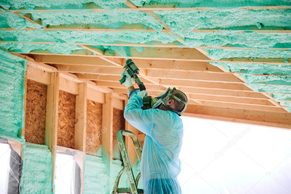 Male builder insulating wooden frame house. Man worker spraying polyurethane foam inside of future cottage, using plural component gun. Construction and insulation concept.
