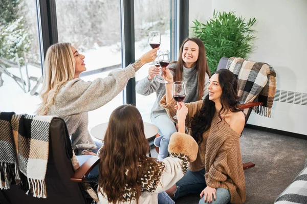 Young women enjoying winter weekends inside contemporary barn house. Four girls having fun and clinking glasses with red wine.