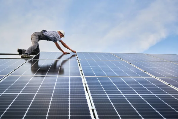 Male worker mounting photovoltaic solar panel system outdoors. Man installer placing solar module on metal rails, wearing construction helmet. Renewable and ecological energy.
