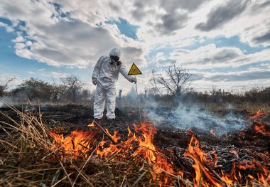 Fireman ecologist extinguishing fire in field. Man in suit and gas mask near burning grass with smoke, holding yellow triangle with skull and crossbones warning sign. Natural disaster concept.
