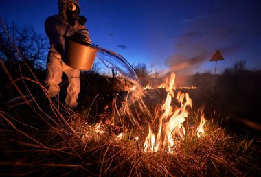 Araştırma bilimadamı, arka planda mavi gökyüzü olan yangını söndürüyor. Koruyucu radyasyon kıyafeti giyen ve gaz maskeli bir adam kovayı tutuyor ve yanan çimlerin üzerine dumanla su döküyor..