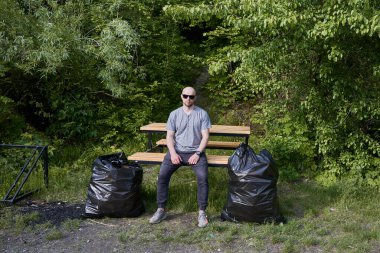 Güneş gözlüklü genç adam çöp torbalarının yanındaki ahşap bankta oturuyor. Yeşil ağaçlar ve çöp torbalarıyla ormanda dinlenen bir erkek. Ekoloji, çöp sorunu ve çevre kirliliği kavramı.