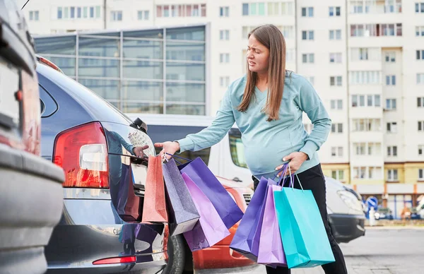 Young pregnant woman with paper bags opening transport trunk at city parking feeling birth contractions. Future mother feeling pain in abdomen belly during maternity shopping.