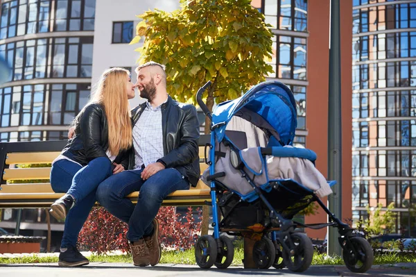 一个快乐的家庭 坐在蓝色的婴儿车里 与一个无法辨认的婴儿坐在一起 享受着 侧视图年轻父母坐在长椅上与婴儿车 背景的现代住宅 — 图库照片