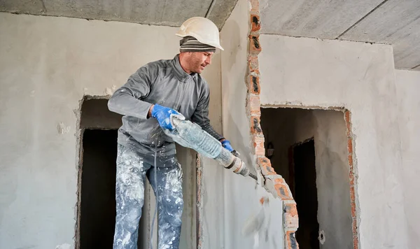 Man builder in construction helmet drilling wall with hammer drill. Male worker using drill breaker while destroying wall in apartment under renovation. Demolition work and home renovation concept.