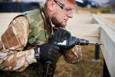 Man building wooden house on pile foundation. Close up of male worker drilling hole by electric drill in wooden frame of future house. Builder wearing protective goggles. Carpentry concept.