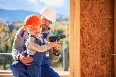 Father with toddler son building wooden frame house. Male builder and kid using screwdriver on construction site, wearing helmet and blue overalls on sunny day. Carpentry and family concept.