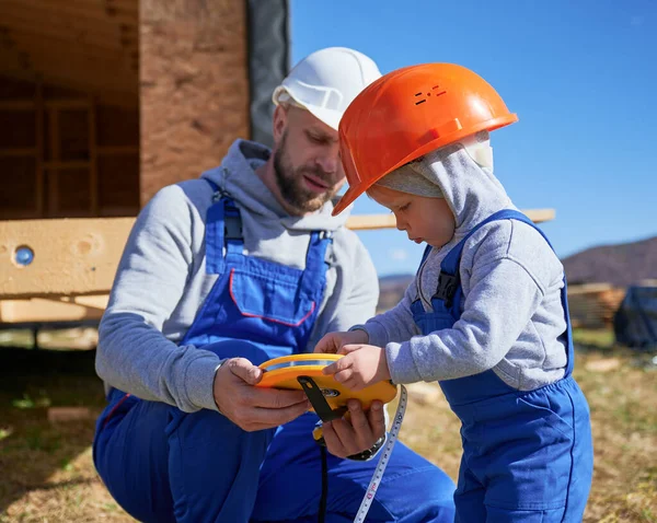 Father with toddler son building wooden frame house. Male builder and kid playing with tape measure on construction site, wearing helmet and blue overalls on sunny day. Carpentry and family concept.