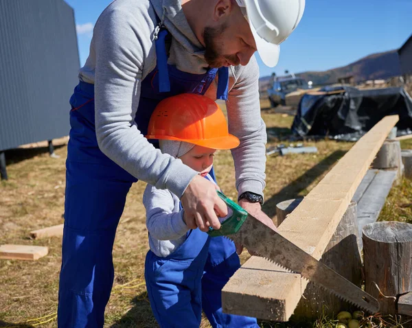 Father with toddler son building wooden frame house. Male builders using hand saw to cut boards on construction site, wearing helmet and blue overalls on sunny day. Carpentry and family concept.