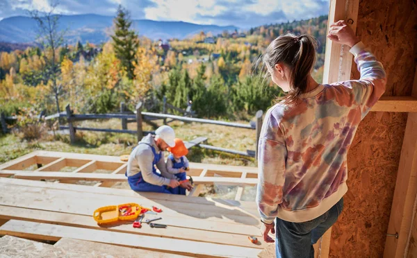 Father, mother and son building wooden frame house. Toddler boy helping his daddy, while woman looking for them on construction site. Guys wearing helmet and blue overalls. Carpentry, family concept.