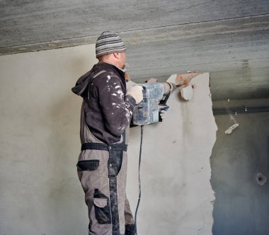 Rear view of professional builder in hat and workwear destroying upper third of wall in the room with jackhammer. Overhaul, redevelopment of apartment space.