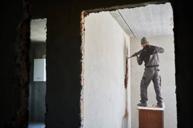 Side view of dismantler in working form working with jackhammer inside apartment. Builder standing on table and carrying out gradual destruction of wall separating two rooms.