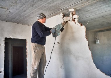 Adult skilled wall demolition specialist destroying inner wall by jackhammer. Overhaul of living space, redevelopment of house. On the background other walls and entrance door.