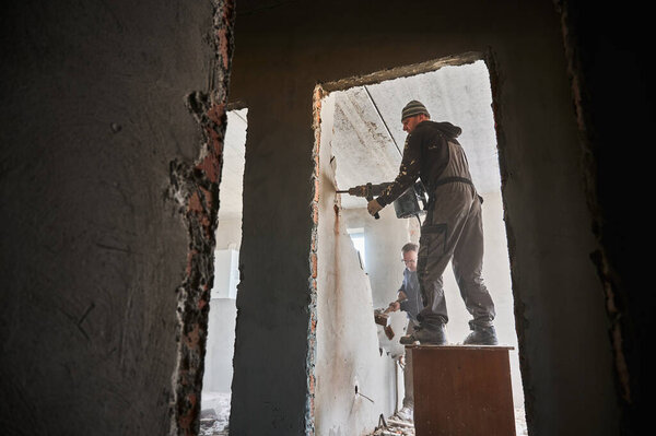 Bottom view of two builders deconstructing interior partition. One worker standing on desk with hammer demolishing upper part of wall, another worker by sledgehammer breaking remaining lower part.