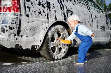 Toddler child leaning over rear wheel of foamy car and holding sponge for washing auto, testing himself in the profession of car washer outdoors in the afternoon.