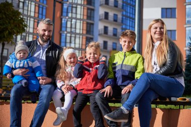 Two adults spending time with cute children, smiling in spring sunny day. Front view of middle-aged parents sitting on bench with four adorable kids, having rest in city centre. Concept of family.