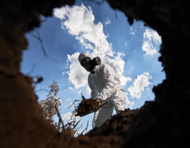 Çevre bilimci, çukur kazıyor, kürek ve ağaç dikiyor, gaz maskesi takıyor ve açık havada koruyucu tulumlar giyiyor, mavi bulutlu gökyüzünün arka planında..