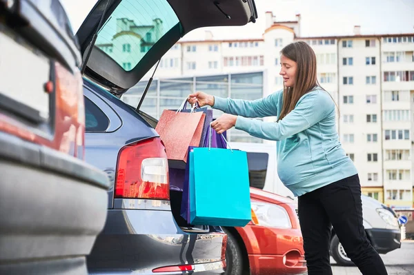 Side view of pregnant woman with belly puting shopping bags in automobile trunk enjoying maternity and parenthood in awaiting mode, pregnant female preparing for baby birth during Black Friday