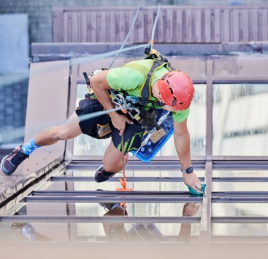Endüstriyel dağcılık işçisi yüksek binaların camlarını yıkıyor, güvenlik halatına asılıyor. Koruyucu başlık takmış, gökdelen cephelerini temizleyen bir adam. Üst görünüm.
