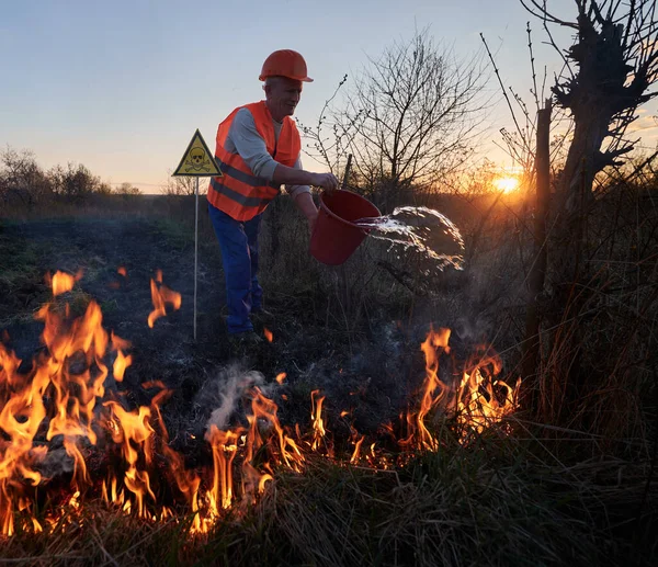 Fireman ecologist extinguishing fire in field in the evening. Male environmentalist holding bucket and pouring water on burning dry grass near yellow triangle with skull and crossbones warning sign.