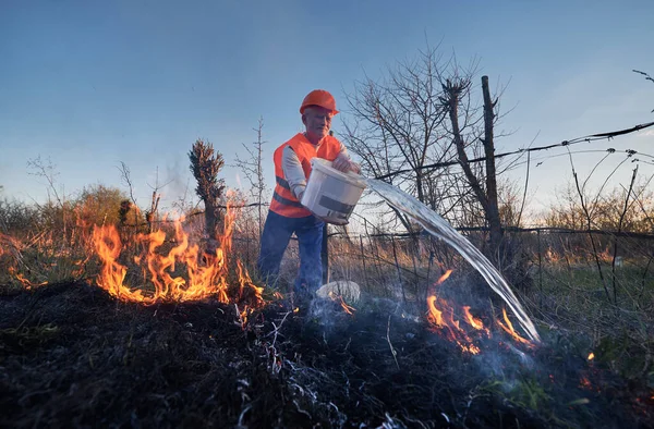 Fireman ecologist extinguishing fire in field with evening sky on background. Male environmentalist holding bucket and pouring water on burning dry grass. Natural disaster concept.