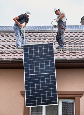 Men workers lifting up photovoltaic solar modul on roof of house. Electricians in helmets installing solar panel system outdoors. Concept of alternative and renewable energy.