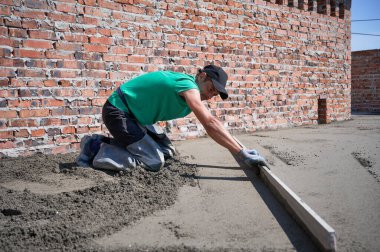 Side view of man in cap placing screed rail on the floor covered with sand-cement mix at construction site. Male worker leveling surface with straight edge while screeding floor in new building.