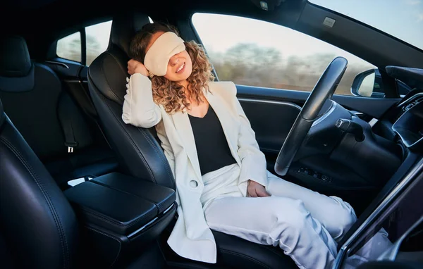 Charming woman in sleeping mask sitting on driver seat and smiling while taking nap in electric car with autopilot system. Businesswoman using driver-assistance system while sleeping in automobile.